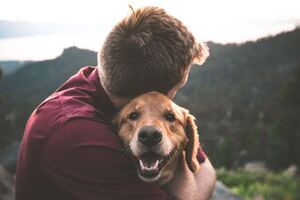 A man is hugging a tan dog in front of mountain trees.