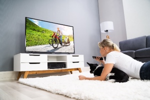 A woman watches TV at home with her dog on a white rug, holding a remote. The concept is that dogs can watch TV.